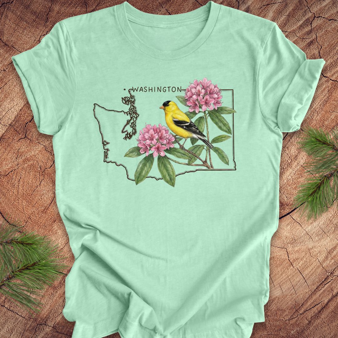 Mint green t-shirt with Washington state outline with state bird goldfinch and rhododendron on a wood background.
