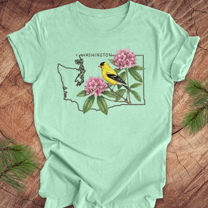 Mint green t-shirt with Washington state outline with state bird goldfinch and rhododendron on a wood background.
