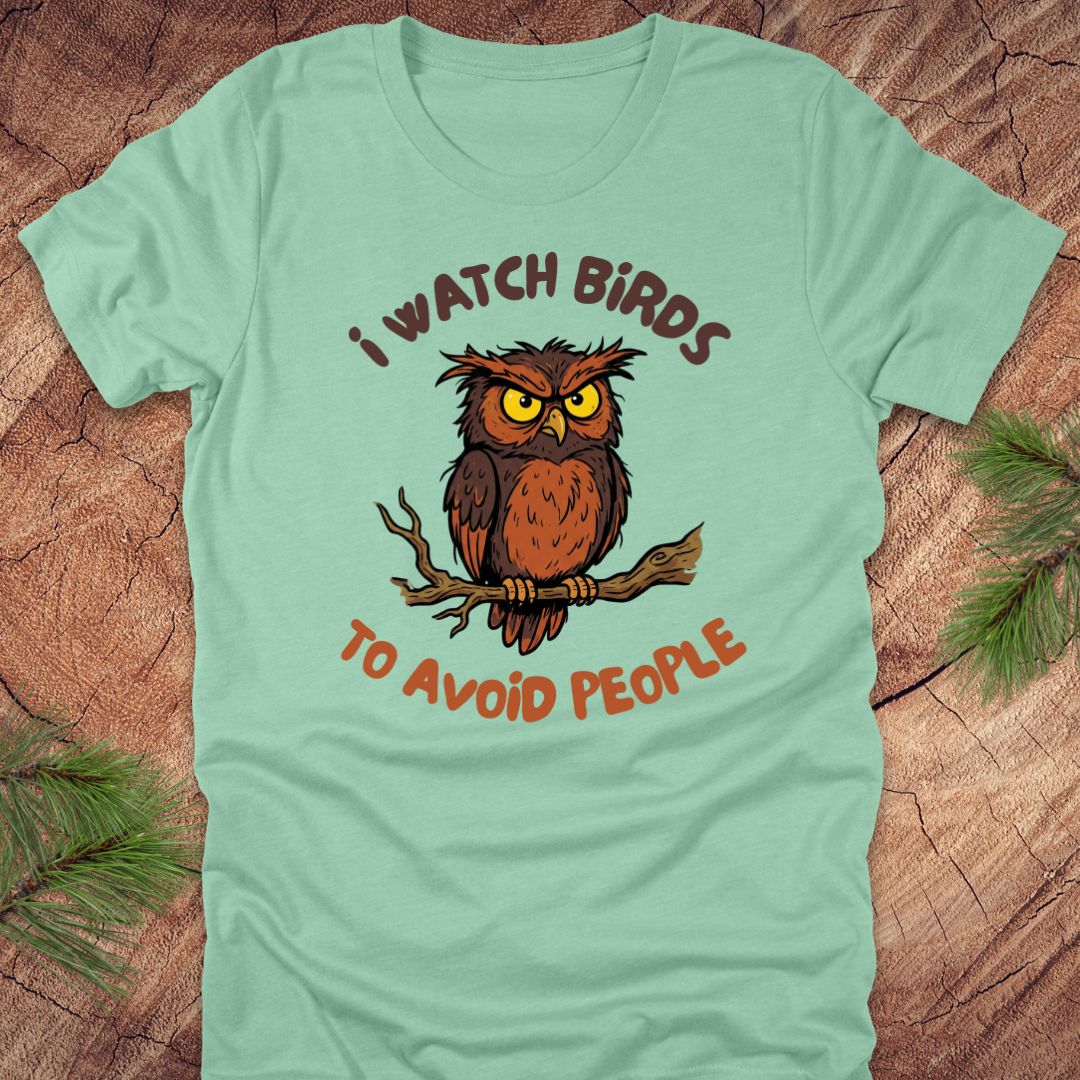Mint green t-shirt with an owl graphic and text, 'I watch birds to avoid people' on a wooden background