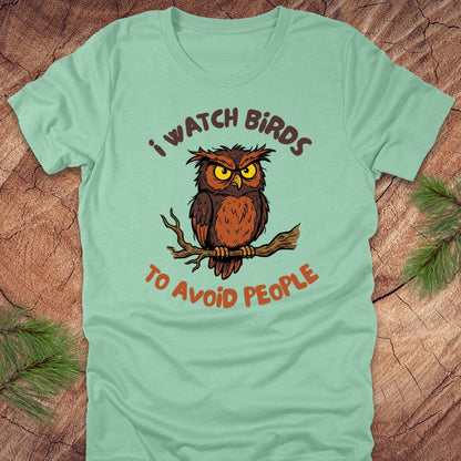 Mint green t-shirt with an owl graphic and text, 'I watch birds to avoid people' on a wooden background