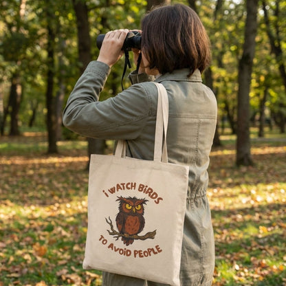 A woman in a park setting with binoculars raised up, holding a canvas tote bag with owl design.