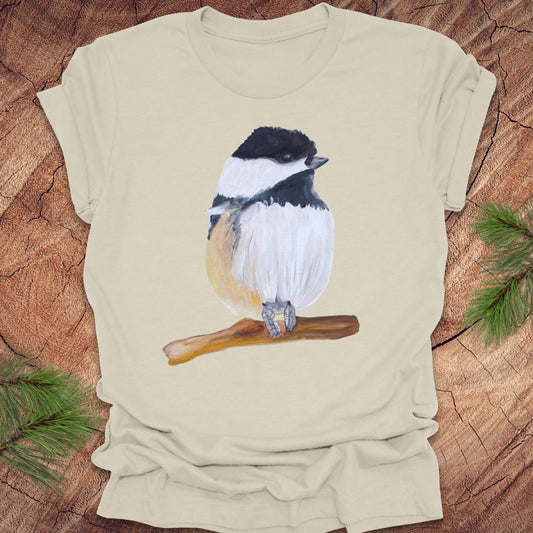 Beige t-shirt with a bird illustration on a wooden surface with pine branches.