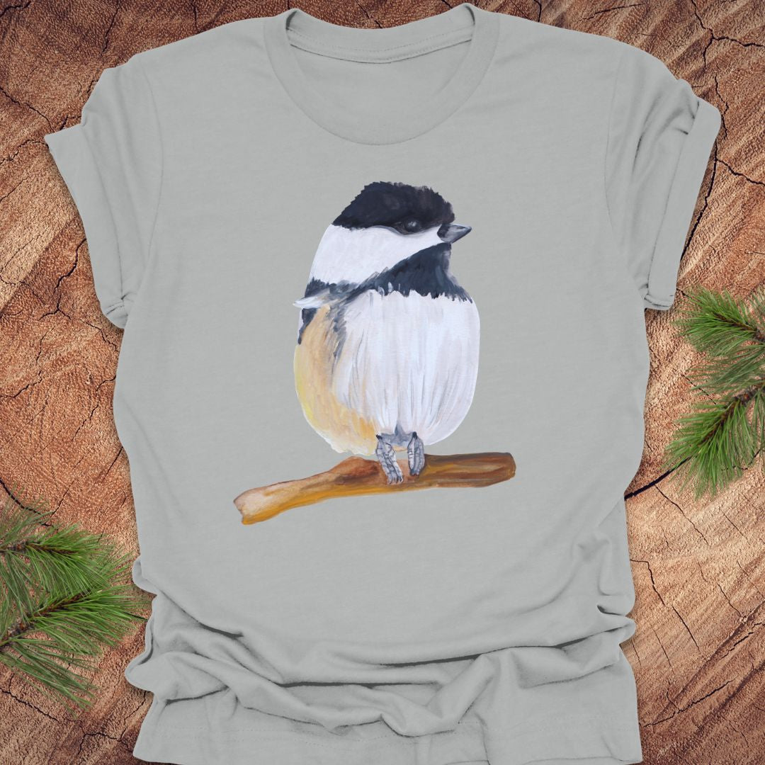 Gray t-shirt with a chickadee on a wooden surface with pine branches.