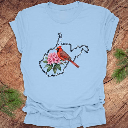 Baby blue t-shirt with the outline of West Virginia and its state bird,and flower cardinal and  rhododendron, on a wooden background.