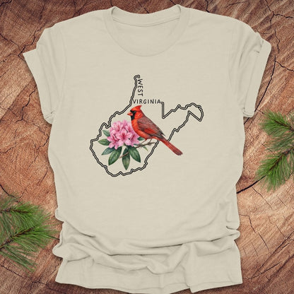 Beige t-shirt with the outline of West Virginia and its state bird,and flower cardinal and  rhododendron, on a wooden background.