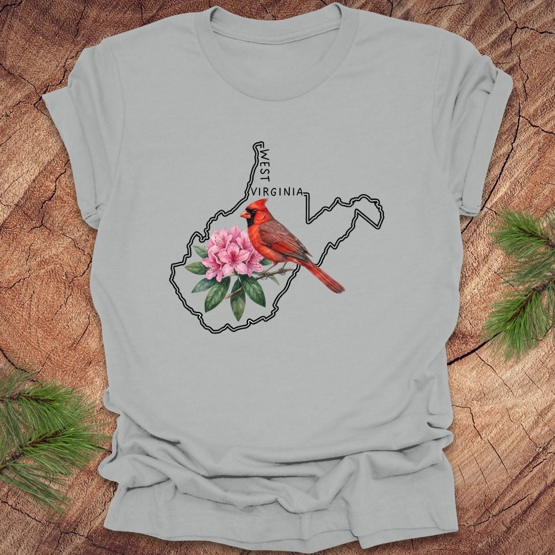 Silver t-shirt with the outline of West Virginia and its state bird,and flower cardinal and  rhododendron, on a wooden background.