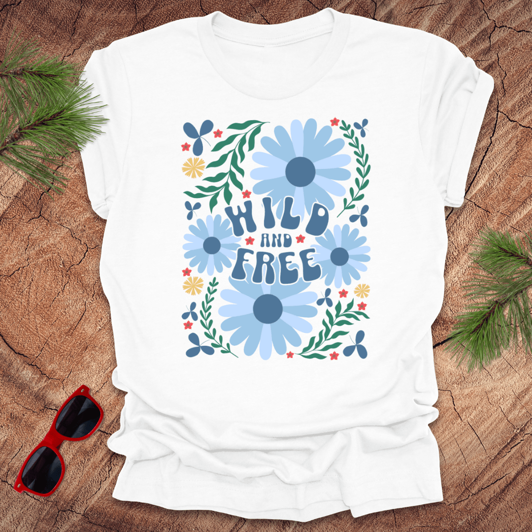 White t-shirt with floral design and 'Wild and Free' text on a wooden surface with sunglasses and greenery.