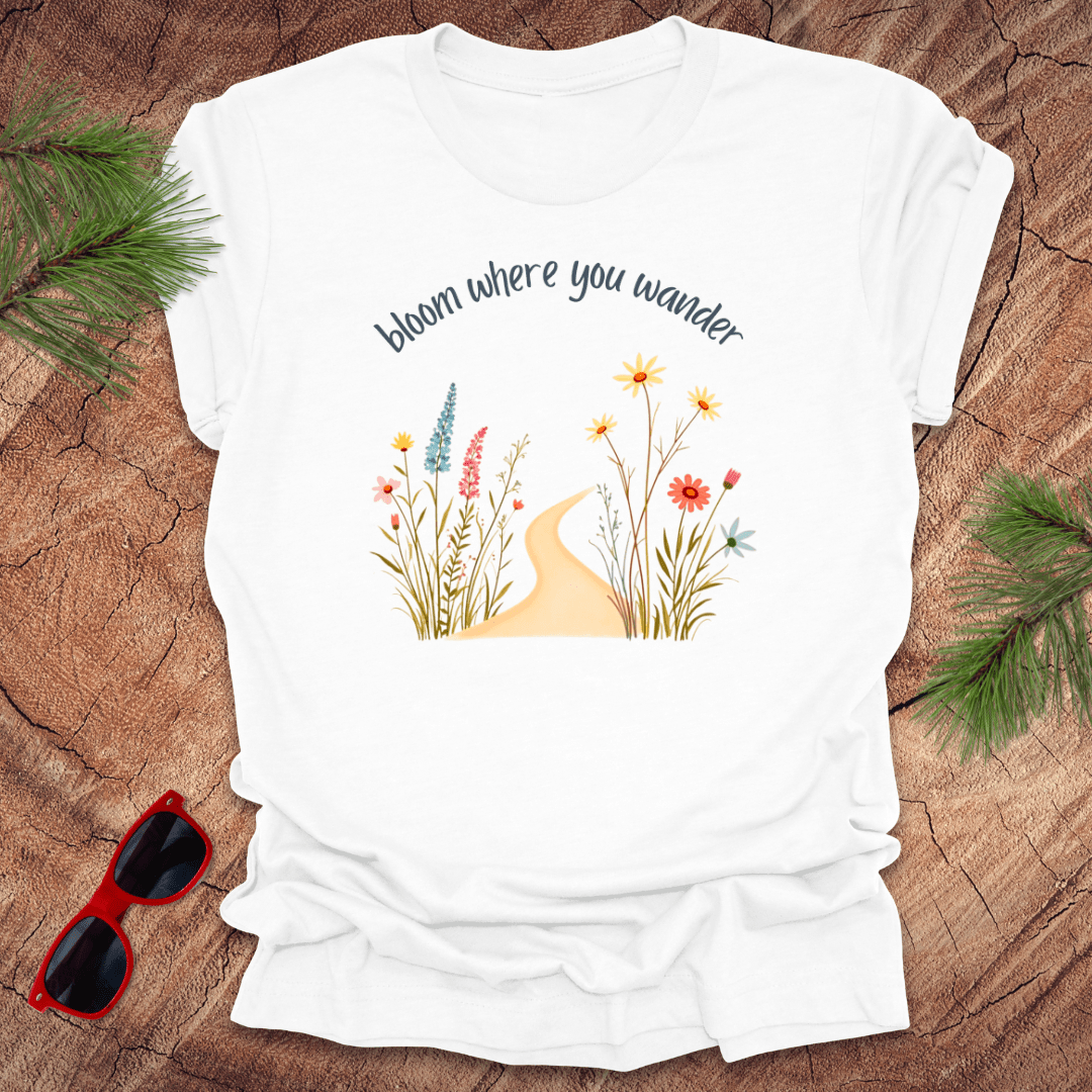 White t-shirt with a graphic that includes a floral design and the text 'bloom where you wander', accompanied by a illustration of flowers on a wooden background with pine needles and red sunglasses to the side.