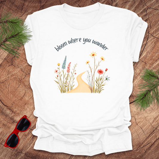 White t-shirt with a graphic that includes a floral design and the text 'bloom where you wander', accompanied by a illustration of flowers on a wooden background with pine needles and red sunglasses to the side.