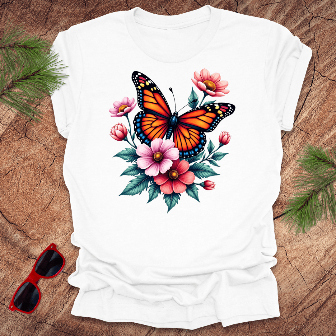 A white t-shirt with a vibrant print of a butterfly surrounded by pink and red flowers, displayed on a wooden surface with green leaves and red sunglasses to the side.