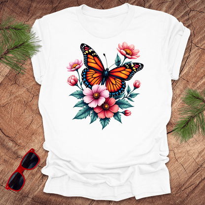 A white t-shirt with a vibrant print of a butterfly surrounded by pink and red flowers, displayed on a wooden surface with green leaves and red sunglasses to the side.