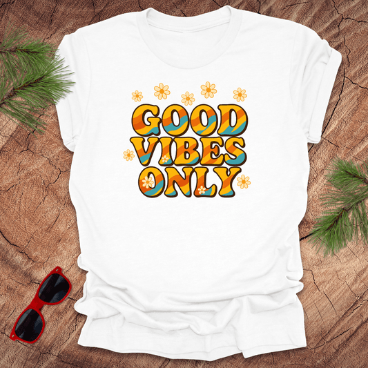 White t-shirt with 'Good Vibes Only' text on a wooden surface with sunglasses and greenery.