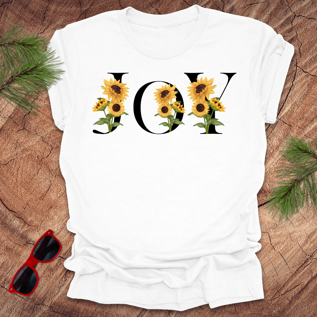 White t-shirt with 'JOY' and sunflower design on a wooden surface with sunglasses and greenery.