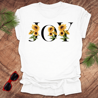 White t-shirt with 'JOY' and sunflower design on a wooden surface with sunglasses and greenery.