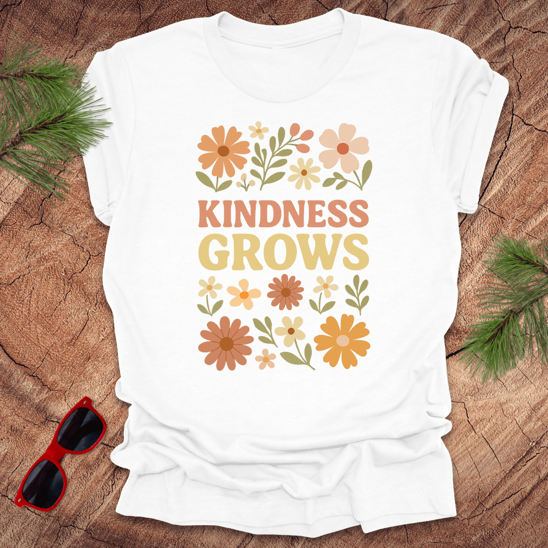 White t-shirt with flowers and text 'Kindness Grows' on a wood background.
