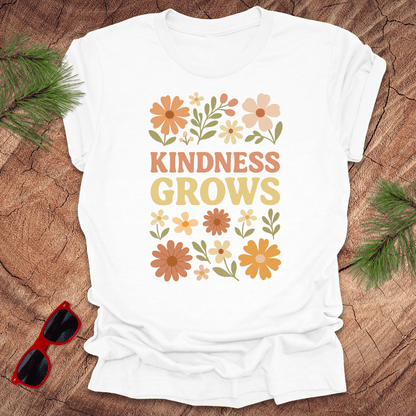 White t-shirt with flowers and text 'Kindness Grows' on a wood background.
