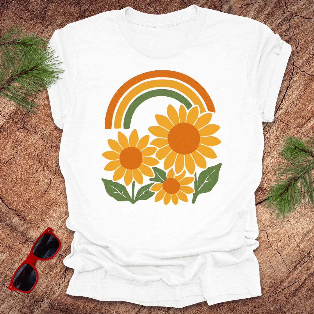 White t-shirt with a rainbow and sunflower design on a wooden surface with sunglasses and greenery.