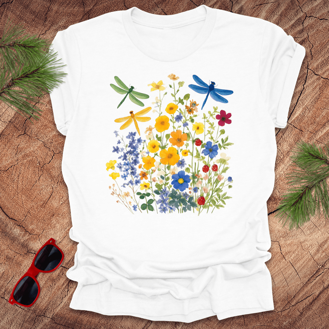 White t-shirt with floral design and dragonflies on a wooden surface with sunglasses and greenery.