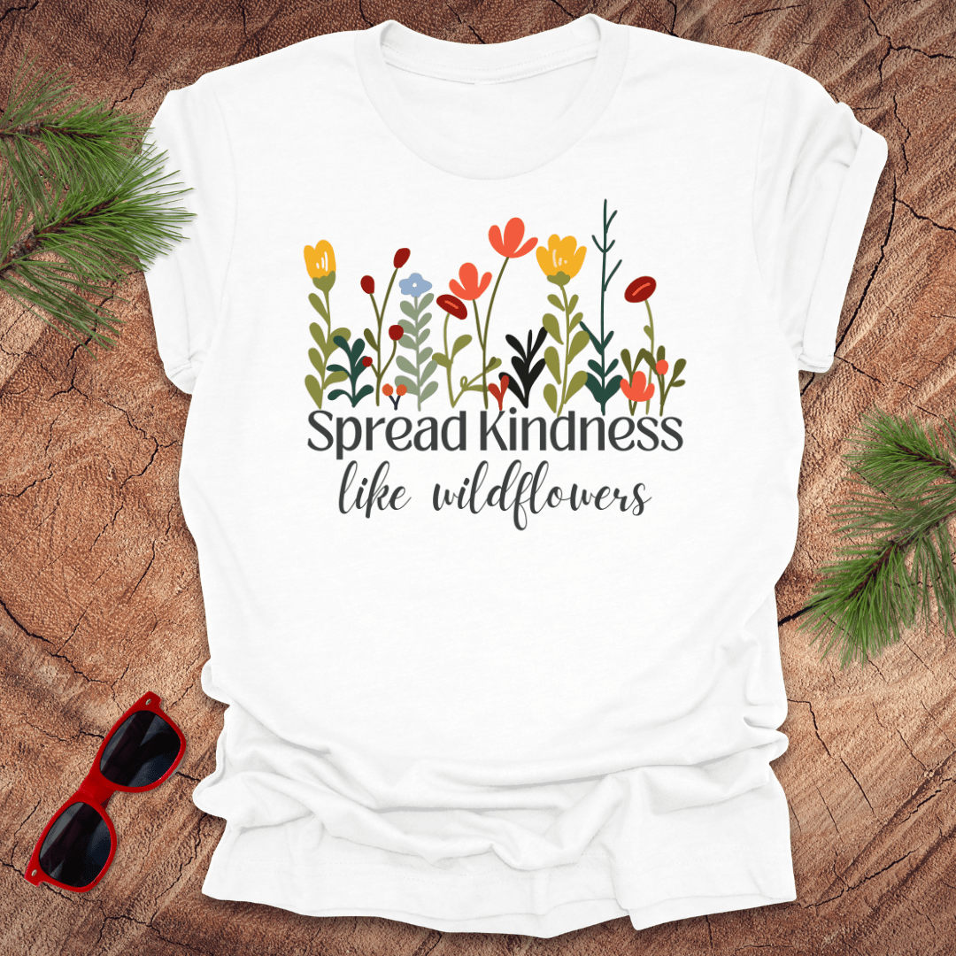 White t-shirt with floral design and 'Spread kindness like wildflowers' text on a wooden surface with sunglasses and greenery.