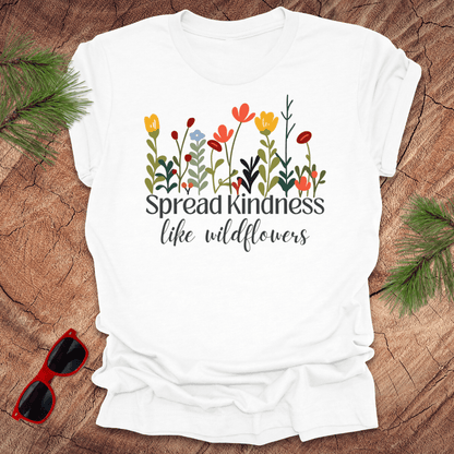White t-shirt with floral design and 'Spread kindness like wildflowers' text on a wooden surface with sunglasses and greenery.