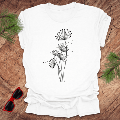 White t-shirt with black floral dandelion design on a wooden surface with sunglasses and greenery.