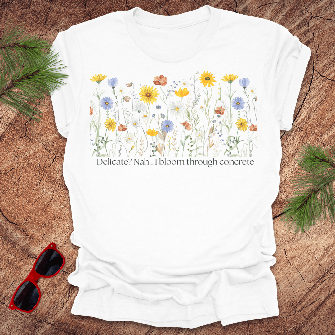 White t-shirt with floral design and text "delicate? nah. i bloom through concrete.' on a wooden surface.
