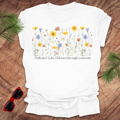 White t-shirt with floral design and text "delicate? nah. i bloom through concrete.' on a wooden surface.