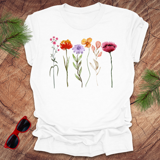 A white t-shirt with a floral design featuring various colors of tall flowers, displayed on a wooden surface with green leaves and red sunglasses to the side.