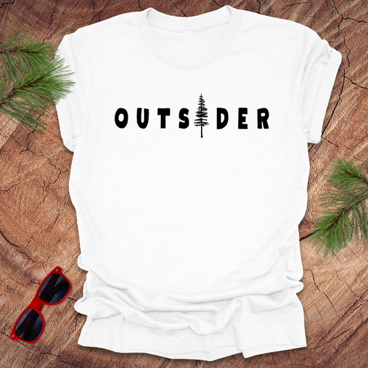 White t-shirt with text 'Outsider" the i is a tree, on a wood background with pine branches and sunglasses.