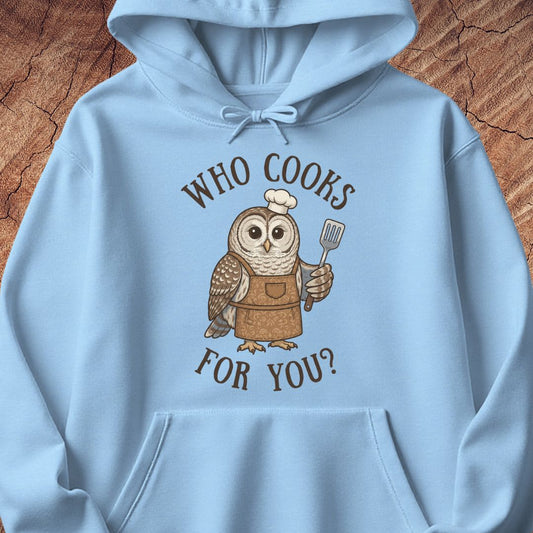 A light blue pullover hoodie featuring a cartoon owl wearing a brown apron and chef's hat, holding a spatula, with the text 'WHO COOKS FOR YOU?' in brown above and below. The hoodie is laid flat on a textured wood background.