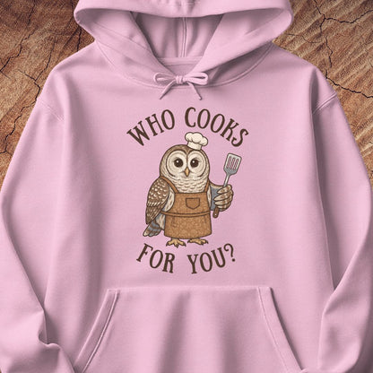 A light pink pullover hoodie featuring a cartoon owl wearing a brown apron and chef's hat, holding a spatula, with the text 'WHO COOKS FOR YOU?' in brown above and below. The hoodie is laid flat on a textured wood background.