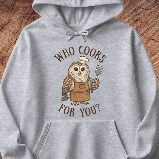 A light heather gray pullover hoodie featuring a cartoon owl wearing a brown apron and chef's hat, holding a spatula, with the text 'WHO COOKS FOR YOU?' in brown above and below. The hoodie is laid flat on a textured wood background.