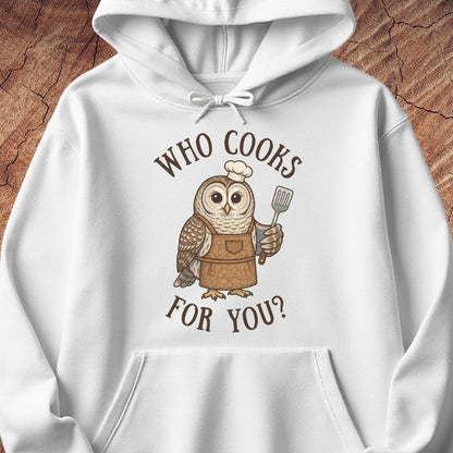 A white pullover hoodie featuring a cartoon owl wearing a brown apron and chef's hat, holding a spatula, with the text 'WHO COOKS FOR YOU?' in brown above and below. The hoodie is laid flat on a textured wood background.
