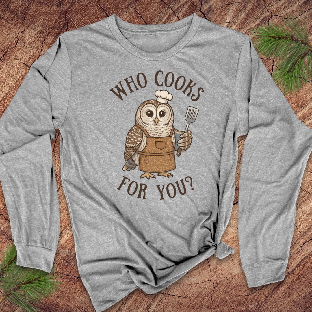A heather gray long-sleeve t-shirt with a graphic of an owl wearing a chef's hat and apron, holding a spatula, with the text 'WHO COOKS FOR YOU?' printed around it. The shirt is laid flat on a rustic wood background with pine branches in the corners.