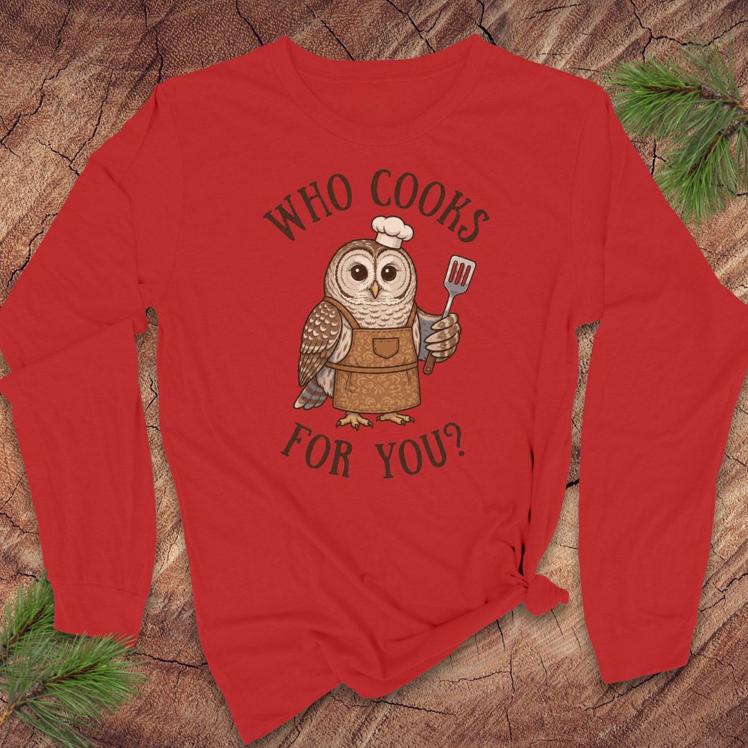 A red long-sleeve t-shirt with a graphic of an owl wearing an apron and holding a spatula, with the text 'WHO COOKS FOR YOU?' printed around it. The shirt is laid flat on a rustic wood background with pine branches in the corners.