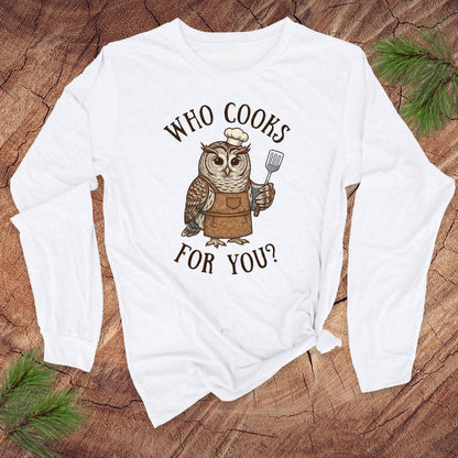 White long-sleeve shirt with an owl graphic and text 'who cooks for you' on a wooden surface