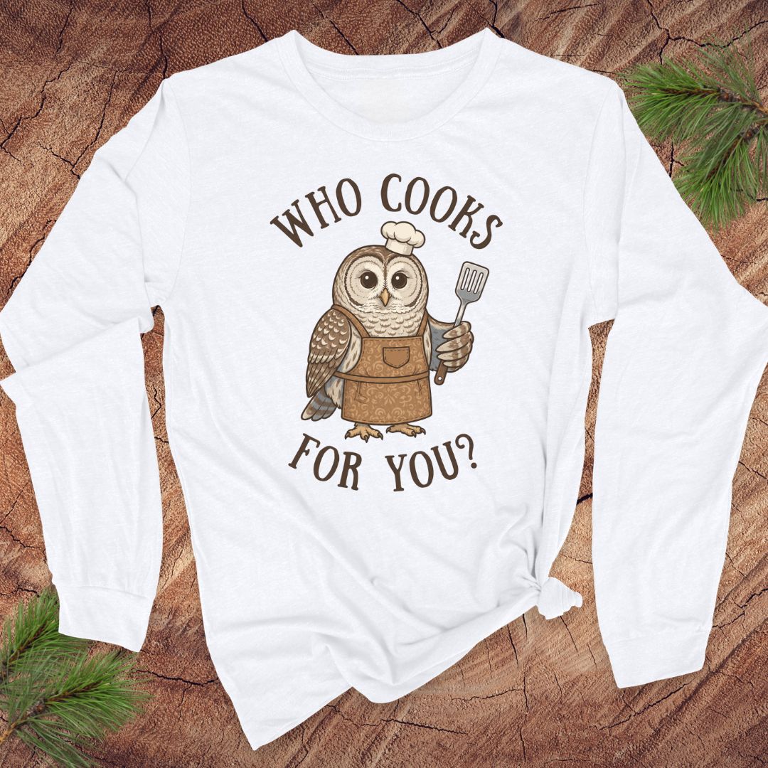 A white long-sleeve t-shirt with a graphic of an owl wearing a chef's hat and apron, holding a spatula, with the text 'WHO COOKS FOR YOU?' printed around it. The shirt is laid flat on a rustic wood background with pine branches in the corners.