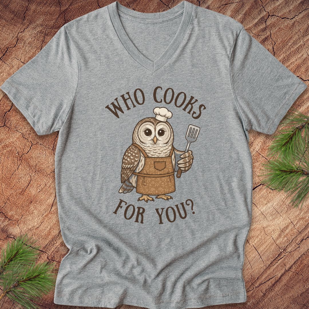 A heather gray V-neck t-shirt featuring a cartoon owl wearing a brown apron and white chef's hat, holding a spatula, with the text 'WHO COOKS FOR YOU?' in brown. The shirt is laid flat on a rustic wood background with pine branches in the corners.