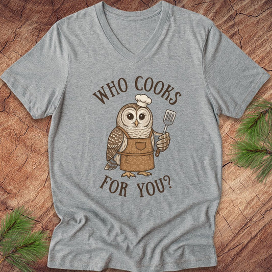 A heather gray V-neck t-shirt featuring a cartoon owl wearing a brown apron and white chef's hat, holding a spatula, with the text 'WHO COOKS FOR YOU?' in brown. The shirt is laid flat on a rustic wood background with pine branches in the corners.