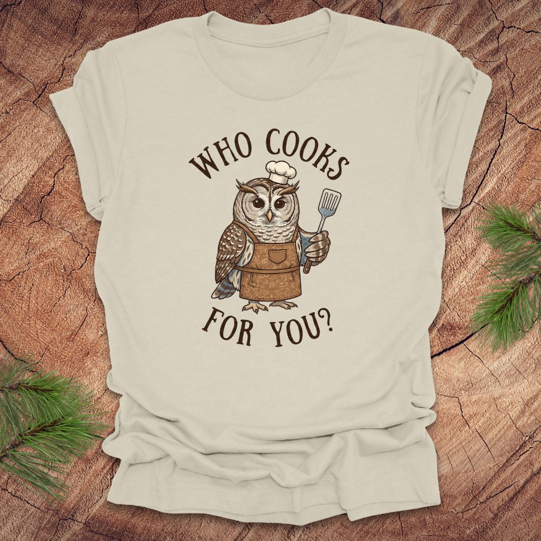 Beige t-shirt with an owl graphic and text 'who cooks for you' on a wooden surface