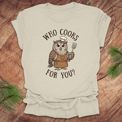 Beige t-shirt with an owl graphic and text 'who cooks for you' on a wooden surface