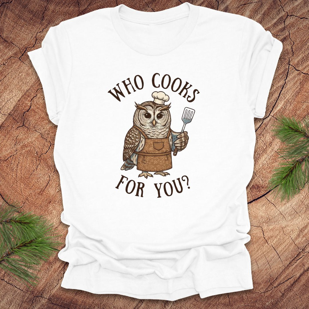 White t-shirt with an owl graphic and text 'who cooks for you' on a wooden surface