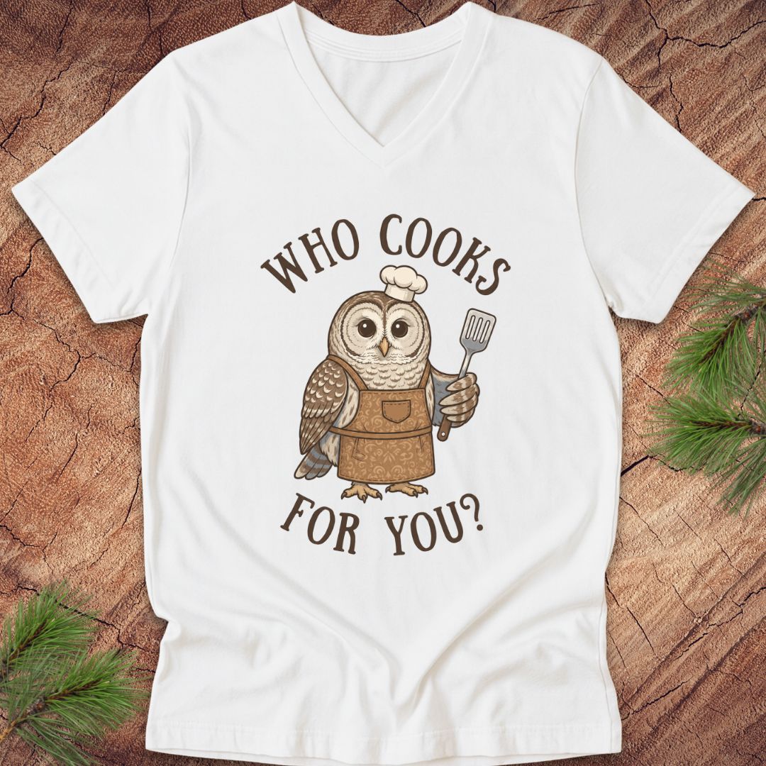 A white V-neck t-shirt laid flat on a rustic wood background with pine branches. The shirt features a graphic of a brown owl wearing a white chef's hat and brown apron, holding a spatula, with the text 'WHO COOKS FOR YOU?' in brown arched lettering.