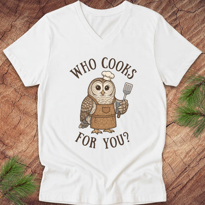 A white V-neck t-shirt laid flat on a rustic wood background with pine branches. The shirt features a graphic of a brown owl wearing a white chef's hat and brown apron, holding a spatula, with the text 'WHO COOKS FOR YOU?' in brown arched lettering.