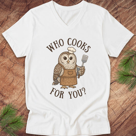 A white V-neck t-shirt laid flat on a rustic wood background with pine branches. The shirt features a graphic of a brown owl wearing a white chef's hat and brown apron, holding a spatula, with the text 'WHO COOKS FOR YOU?' in brown arched lettering.