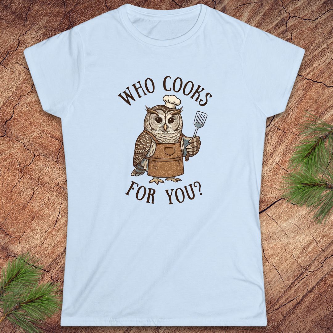 A light blue women's short-sleeve t-shirt with a graphic of a cartoon owl wearing a chef's hat and apron, holding a spatula, with the text 'WHO COOKS FOR YOU?' in brown. The shirt is laid flat on a rustic wood background with pine needles in the corners.