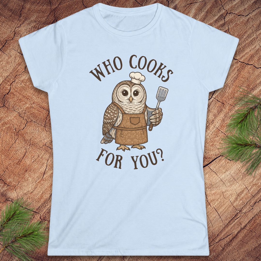 A light blue short-sleeve crewneck t-shirt featuring a cartoon owl wearing a brown apron and chef's hat, holding a spatula, with the text 'WHO COOKS FOR YOU?' in brown. The t-shirt is displayed flat on a rustic wood background with pine needles in the corners.