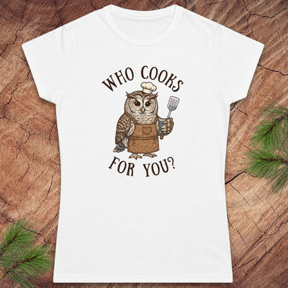 A white short-sleeve crewneck t-shirt featuring a cartoon owl dressed as a chef with the text 'WHO COOKS FOR YOU?' printed on the front, displayed flat on a rustic wood background with pine needles.