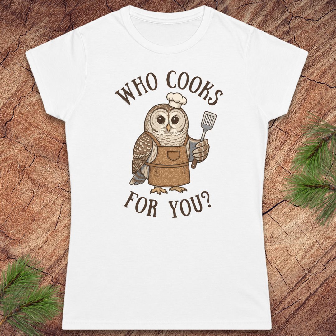A white short-sleeve crewneck t-shirt featuring a cartoon owl wearing a brown apron and chef's hat, holding a spatula, with the text 'WHO COOKS FOR YOU?' printed in brown. The shirt is displayed flat on a textured wood background with pine needles in the corners.