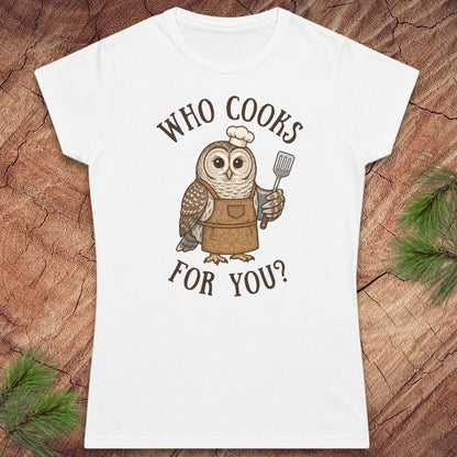 A white short-sleeve crewneck t-shirt featuring a cartoon owl wearing a brown apron and chef's hat, holding a spatula, with the text 'WHO COOKS FOR YOU?' printed in brown. The shirt is displayed flat on a textured wood background with pine needles in the corners.